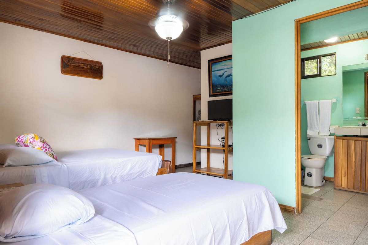 A bedroom with two beds, a wooden ceiling, a television, and a bathroom with a toilet and sink.