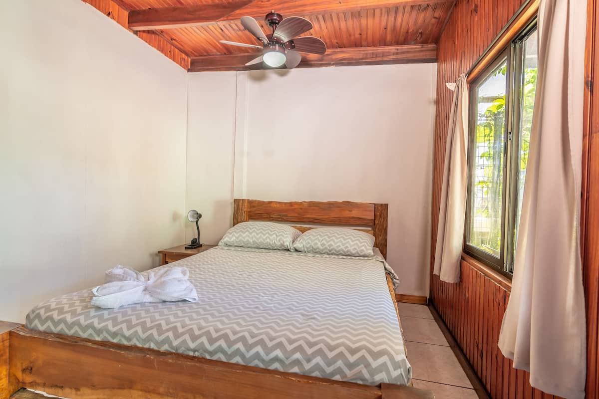 A single bed with a wooden headboard, a ceiling fan, a nightstand with a lamp, and a window with curtains.