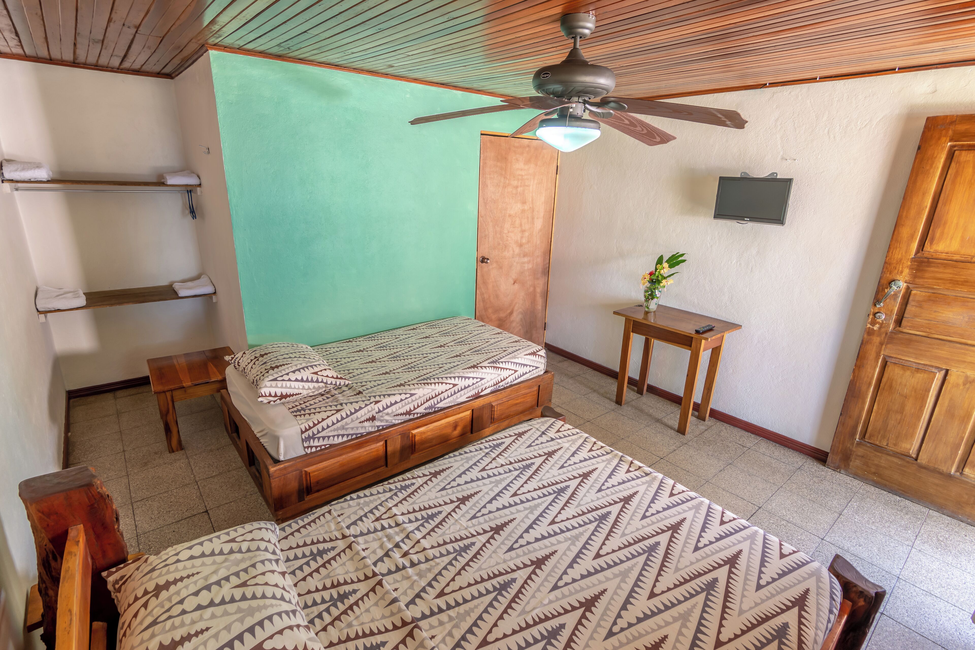 A room with a bed, a wooden ceiling fan, a green wall, a small table with a plant, and a wooden door.