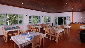 Un espace repas avec des tables et des chaises en bois, de grandes fenêtres et un sol carrelé.