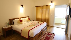 Une chambre d’hôtel avec un grand lit, une tête de lit en bois, deux tables de chevet, une petite table, un tapis à motifs rouges et beiges, une porte-fenêtre coulissante donnant sur le balcon et un téléviseur fixé au mur.