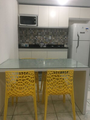 Cozinha com armários brancos, micro-ondas, geladeira e ilha de vidro com duas cadeiras amarelas.