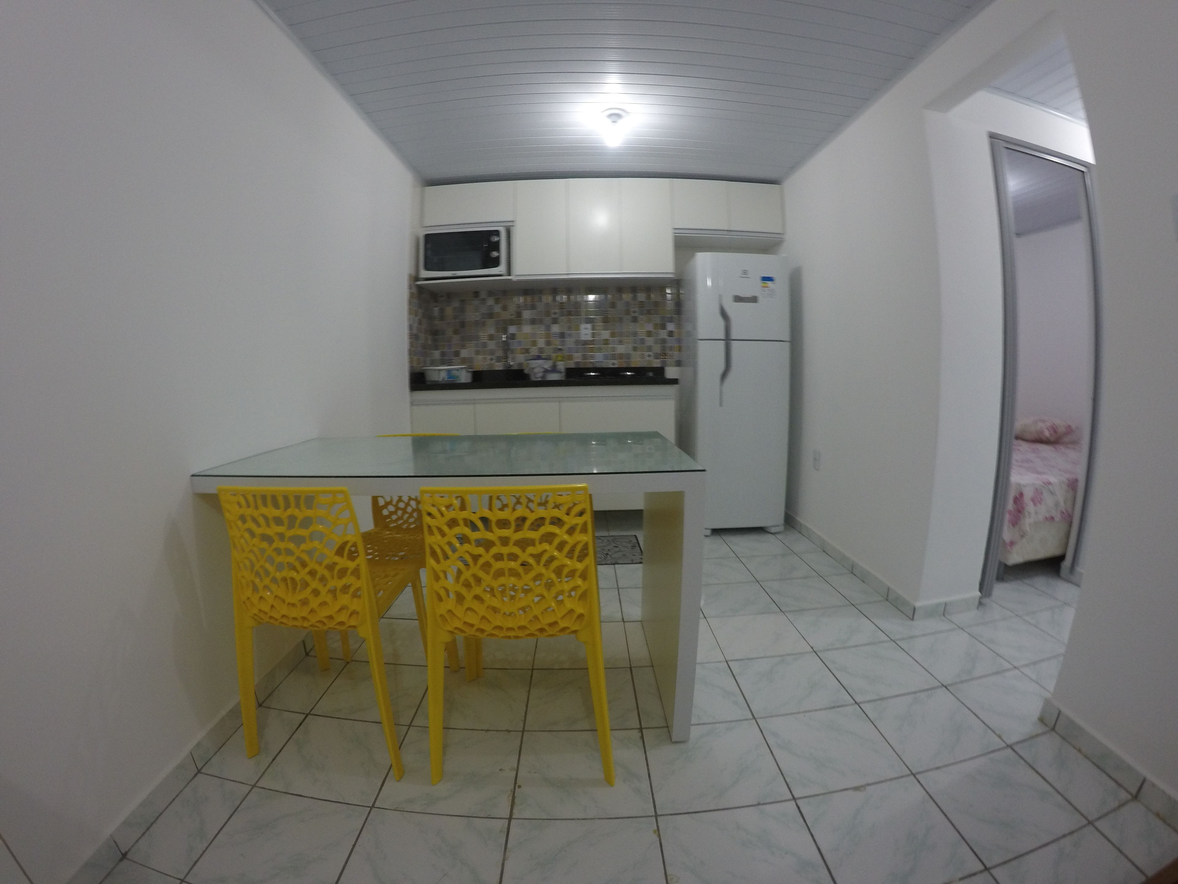 A compact kitchen with a glass-top table and yellow chairs, a microwave, and a refrigerator.