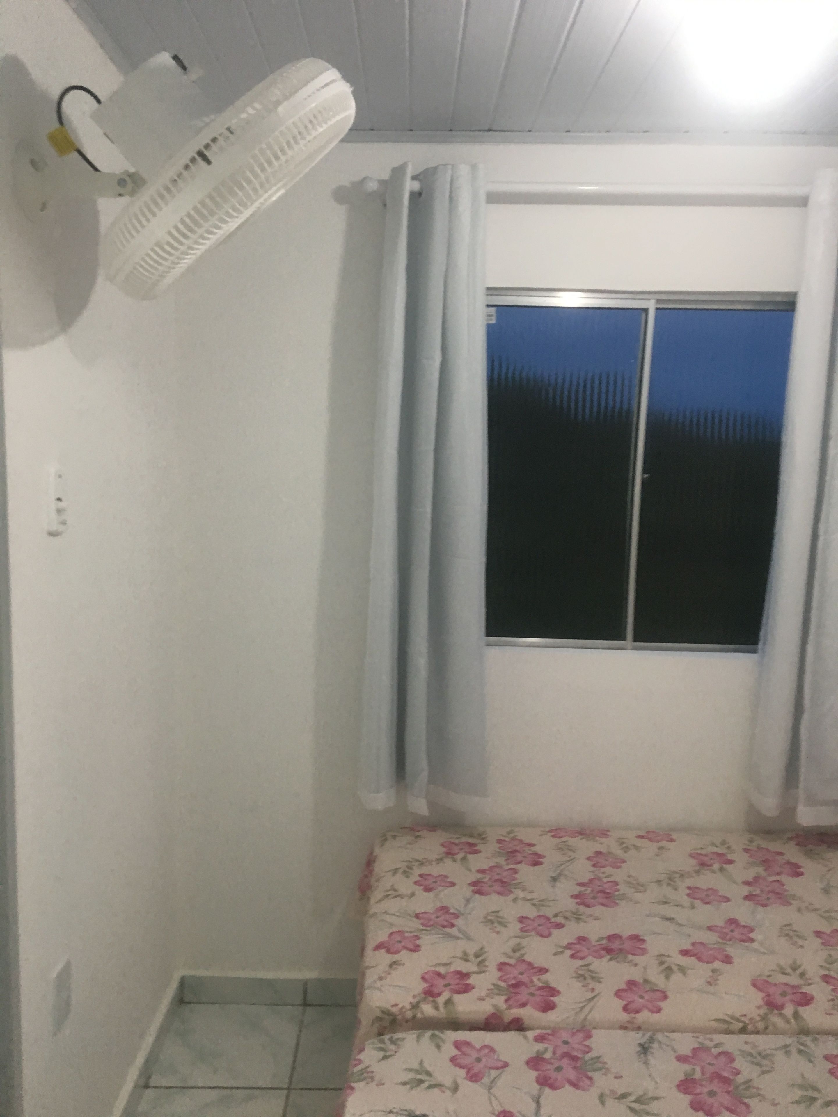 A room with a bed, a window with curtains, a ceiling fan, and a wall-mounted heater.