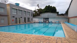 A swimming pool with a tiled deck, surrounded by buildings with a checkered pattern and a sign advertising various amenities. - Pousada Clube Santa Cruz (Santa Cruz de Minas)