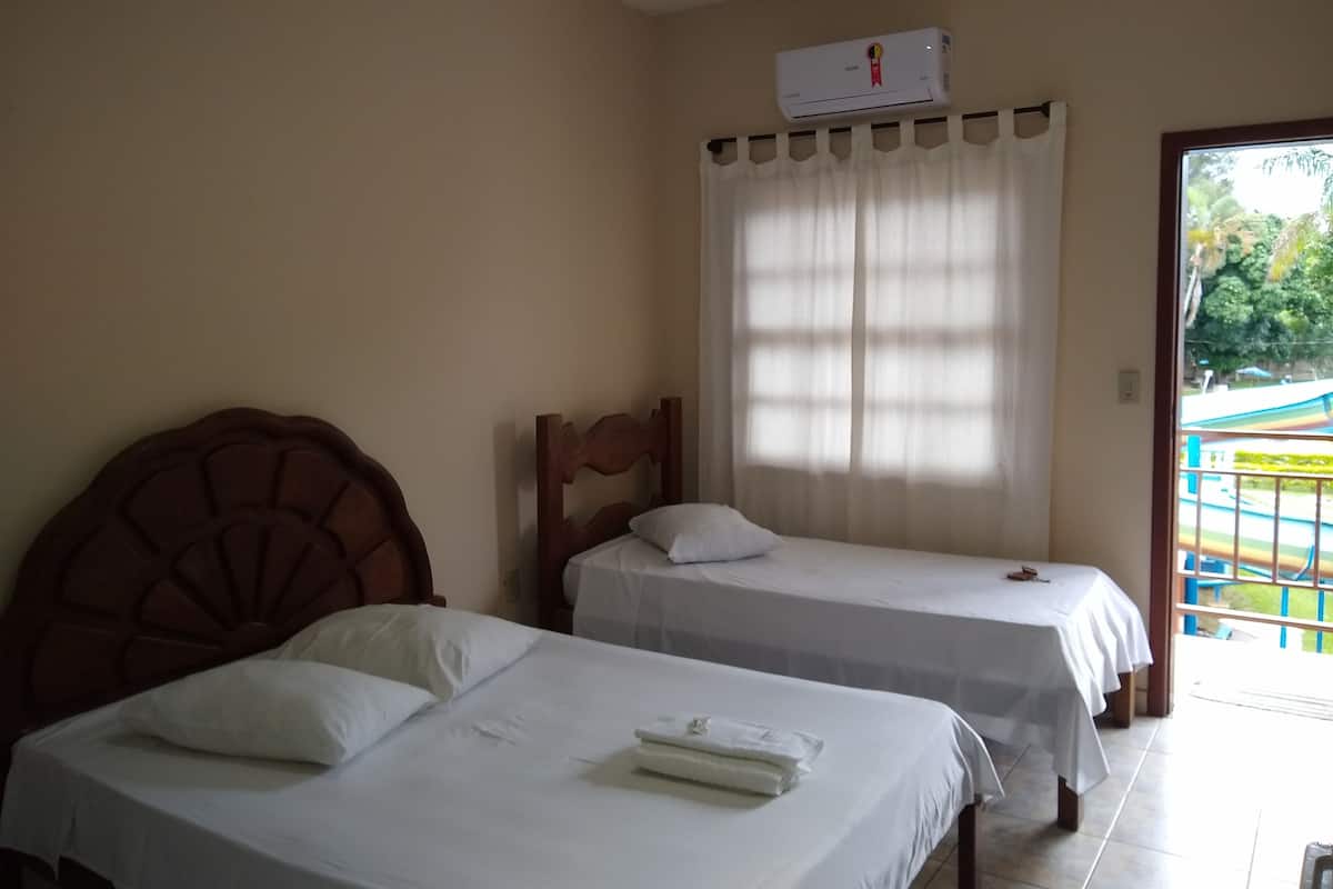 Two single beds with wooden headboards, a white air conditioning unit on the wall, and a view of a colorful outdoor area through a window with white curtains.
