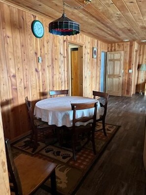 Interior de madeira com uma mesa redonda e cadeiras, um relógio na parede e uma porta ao fundo.