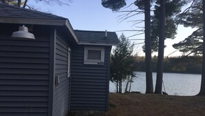 Pieni talo, jossa on ikkuna ja ovi, ja joka sijaitsee vesistön vieressä. Taustalla näkyy puita.