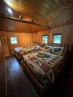 A wooden cabin bedroom with a large bed, two windows, and a wooden ceiling.