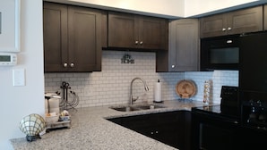A kitchen with dark brown cabinets, white subway tiles, a stainless steel sink, a black refrigerator, and a wooden cutting board with a palm tree design. - Modern Beach Property recently updated (Surfside Beach)