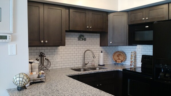 A kitchen with dark brown cabinets, white subway tiles, a stainless steel sink, a black refrigerator, and a wooden cutting board with a palm tree design.