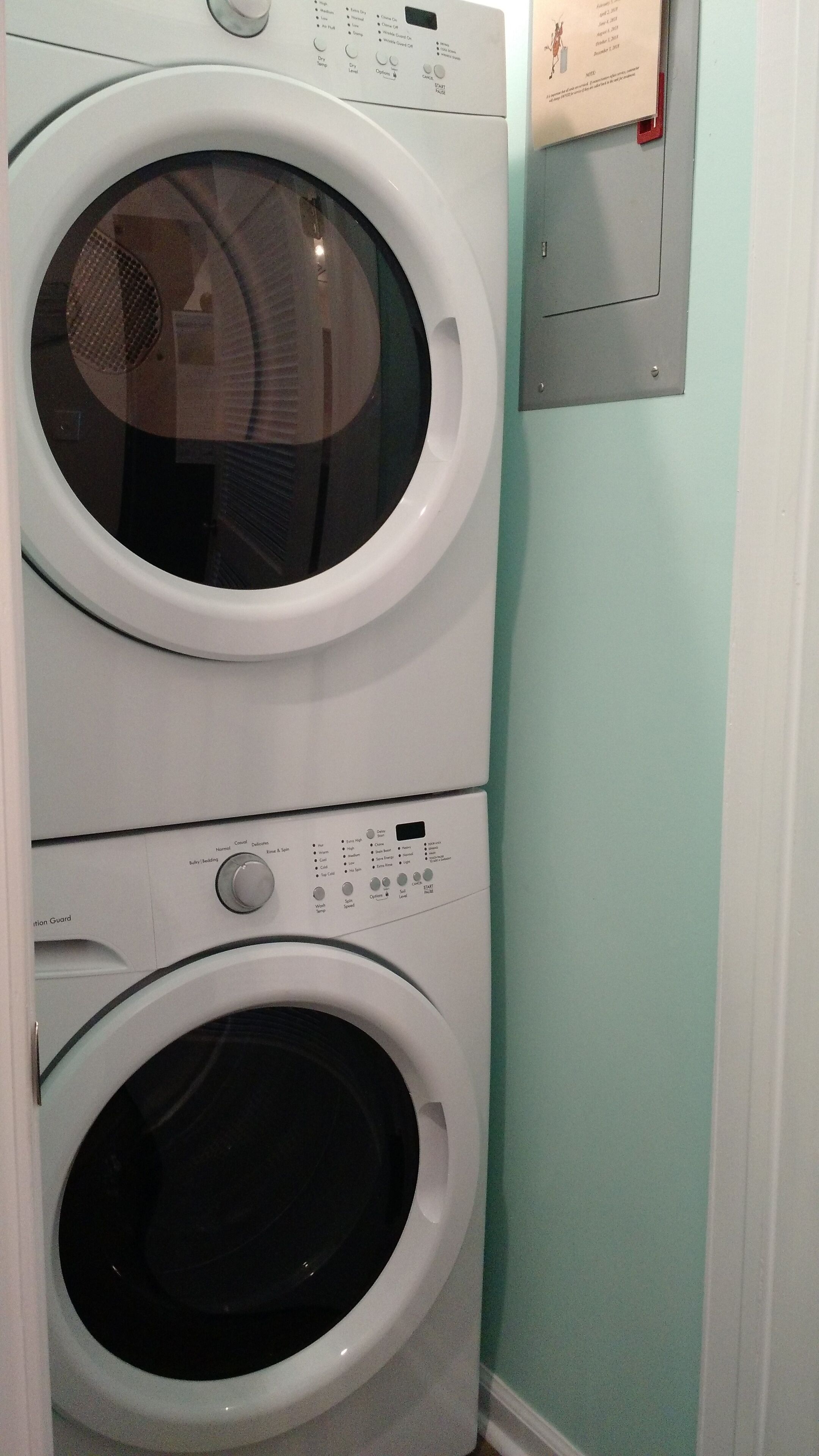 Un appareil combiné lave-linge et sèche-linge, de type « superposé », doté de panneaux de commande et d’une boîte électrique fixée au mur.