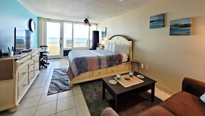 Ein Hotelzimmer mit Bett, Schreibtisch, Stuhl, Kommode, einem kleinen Tisch und Meerblick durch eine Schiebetür.