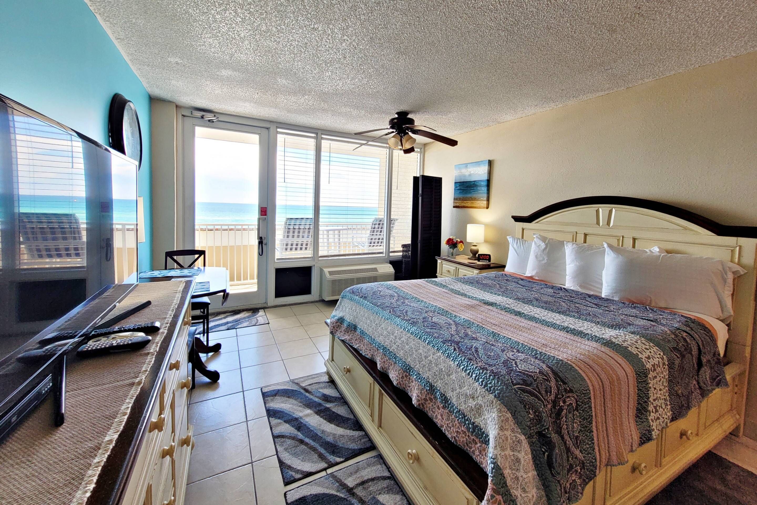 A bedroom with a bed, a desk, a chair, a ceiling fan, and a view of the ocean through large windows.