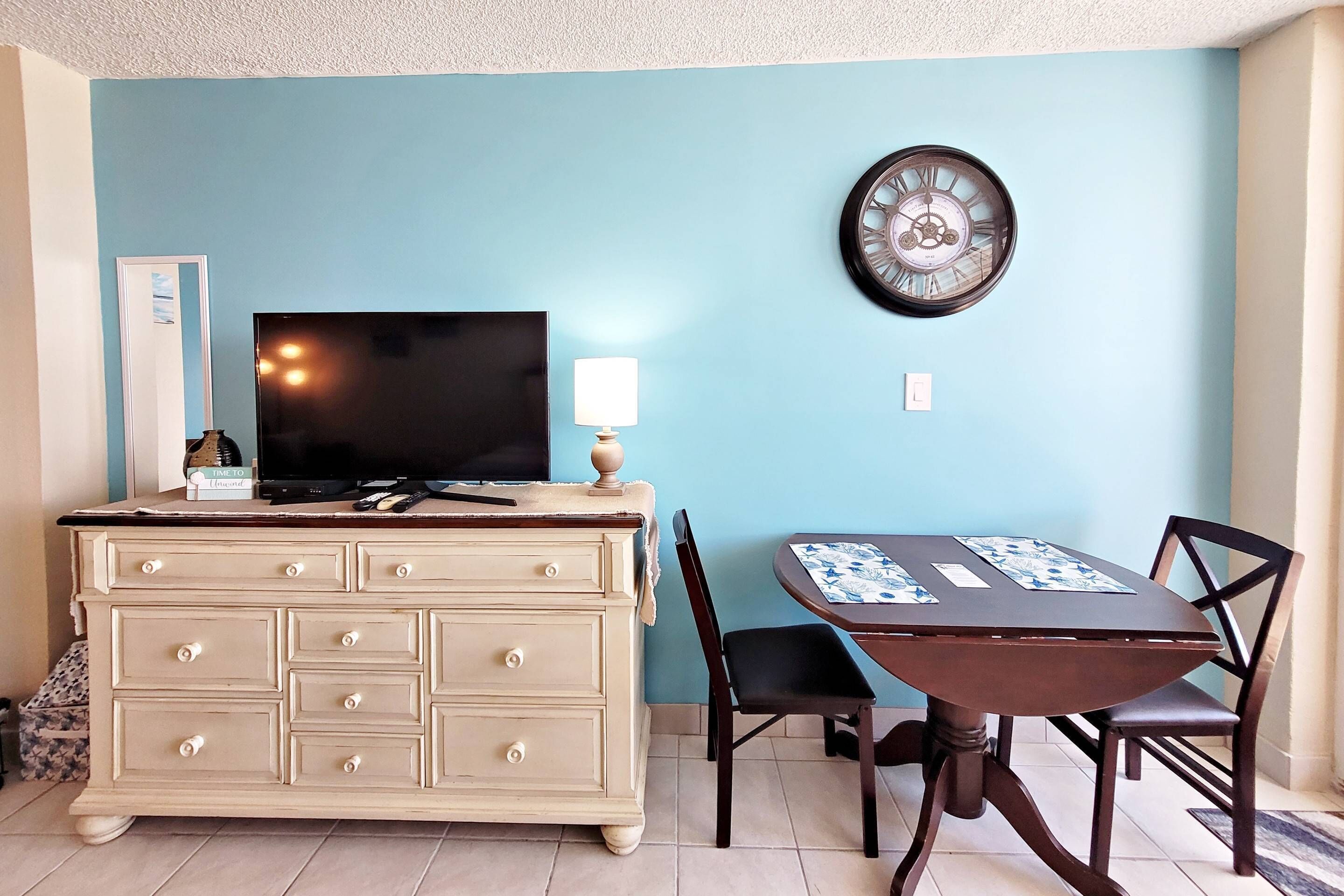 A room with a TV on a dresser, a clock on the wall, and a table with chairs.