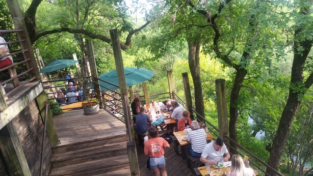 An outdoor wooden deck with tables and chairs, surrounded by trees.