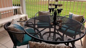 A balcony with a round metal table, two chairs, and a bench. The balcony overlooks a beach with a fence and a sign in the distance. - 🔥 Beautifully Remodeled Grand Beach LOWEST Rates 🦀 Pool & Heated HotTub👙 (Gulf Shores)