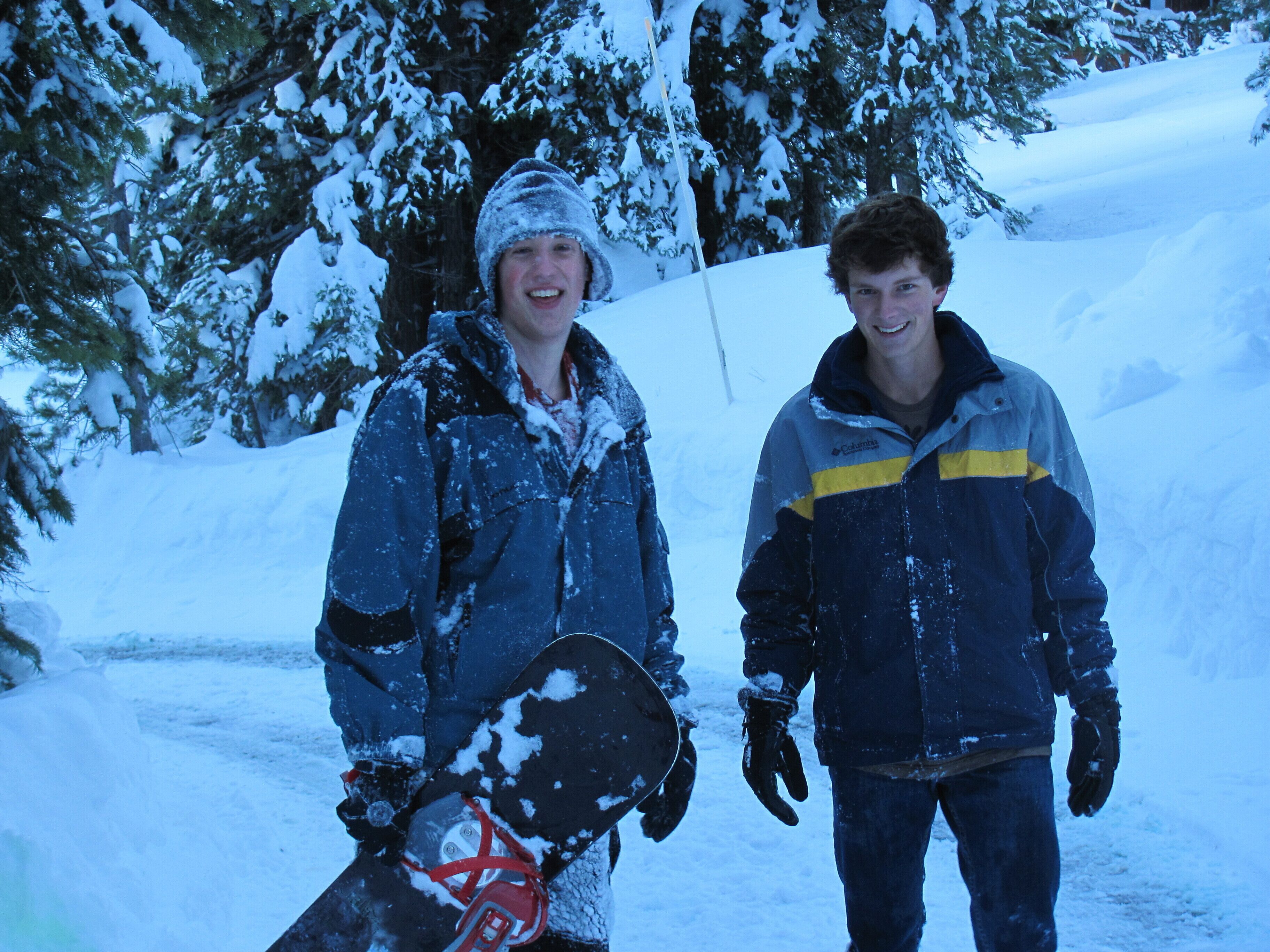 Dos personas con abrigos de invierno, una sosteniendo una tabla de snowboard, en un entorno nevado al aire libre.
