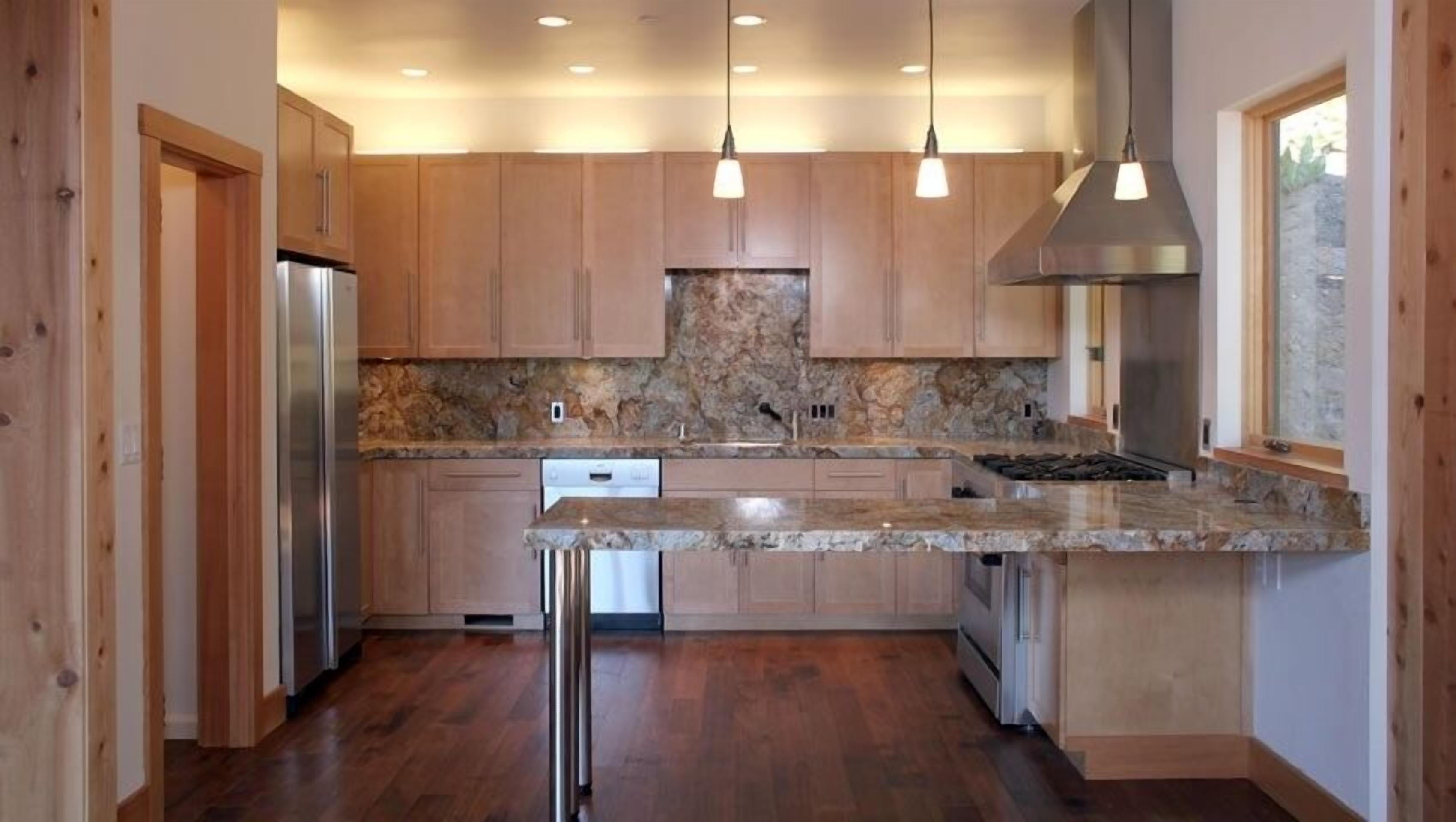 Una cocina moderna con gabinetes de madera, encimeras de granito, electrodomésticos de acero inoxidable e iluminación colgante.