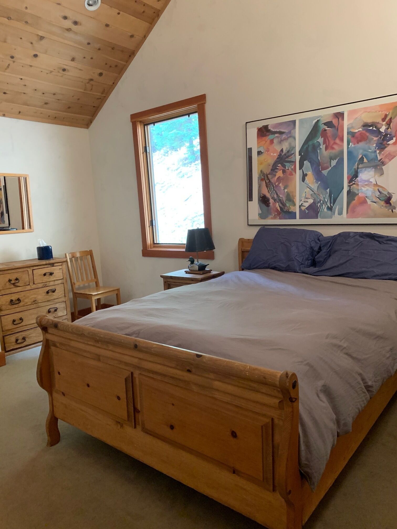 A bedroom with a wooden bed, a dresser, a chair, a nightstand with a lamp, and a window with a view of trees.