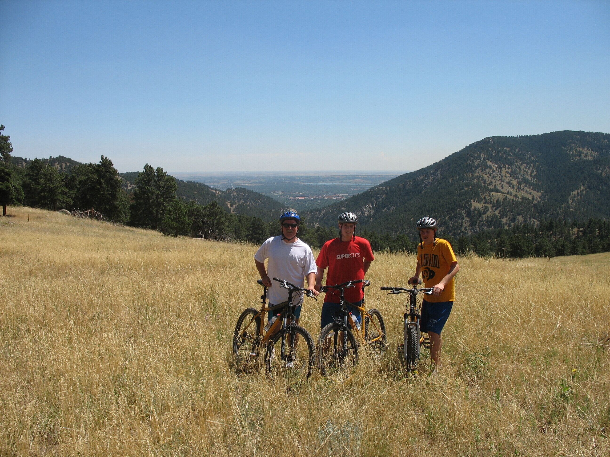 Trois personnes se tiennent sur une colline herbeuse, à côté de leurs vélos, et contemplent une vallée et des montagnes.