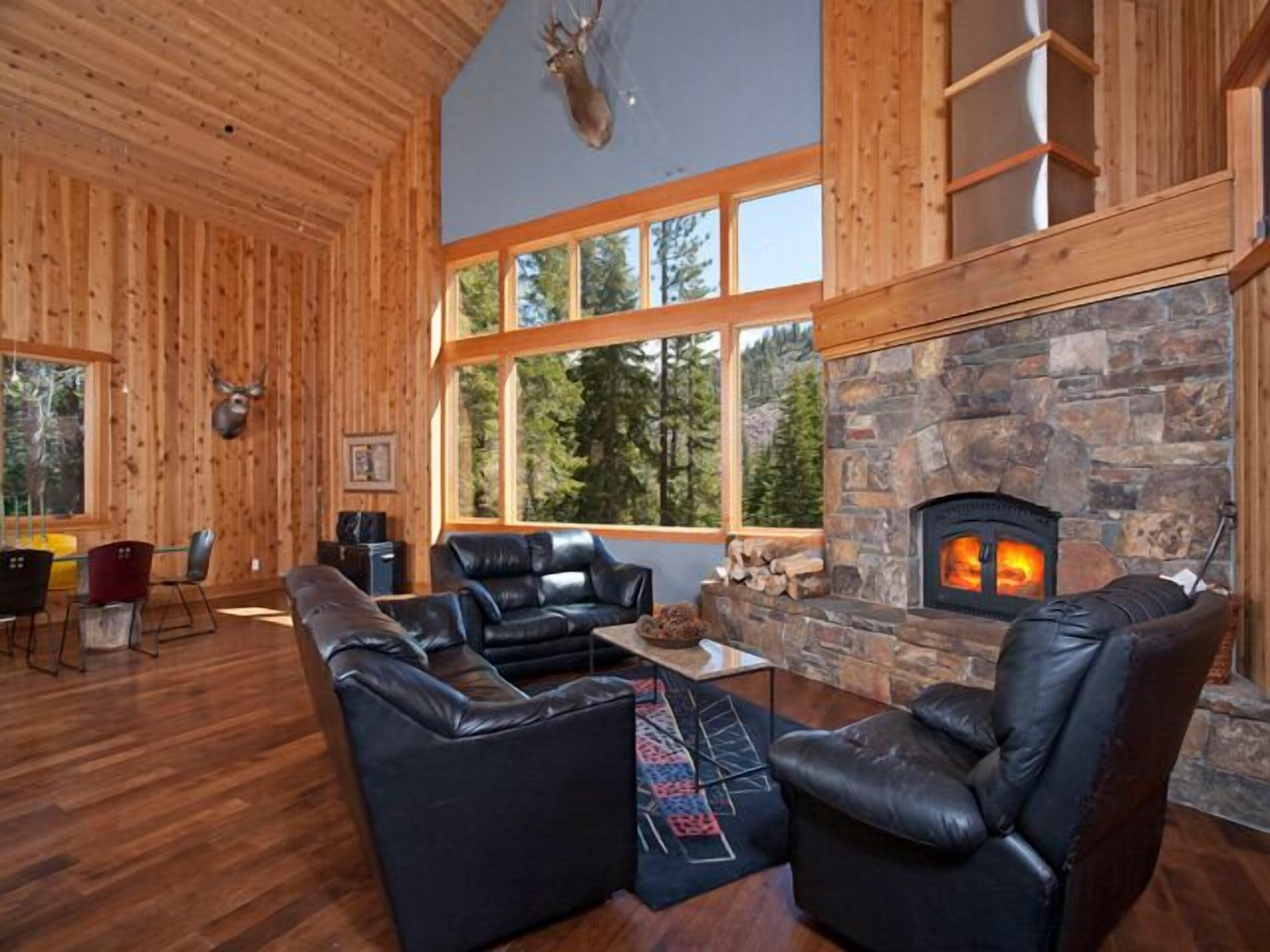 A log cabin interior with a stone fireplace, leather sofas, and large windows offering a forest view.