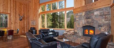 A log cabin interior with a stone fireplace, leather sofas, and large windows offering a forest view.