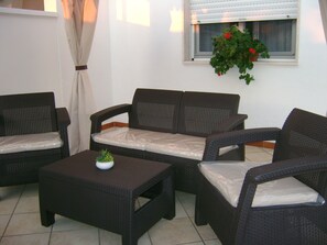 Un ensemble de meubles de jardin en osier comprenant un canapé avec des coussins, une petite table avec une plante et une fenêtre agrémentée d’une plante suspendue.