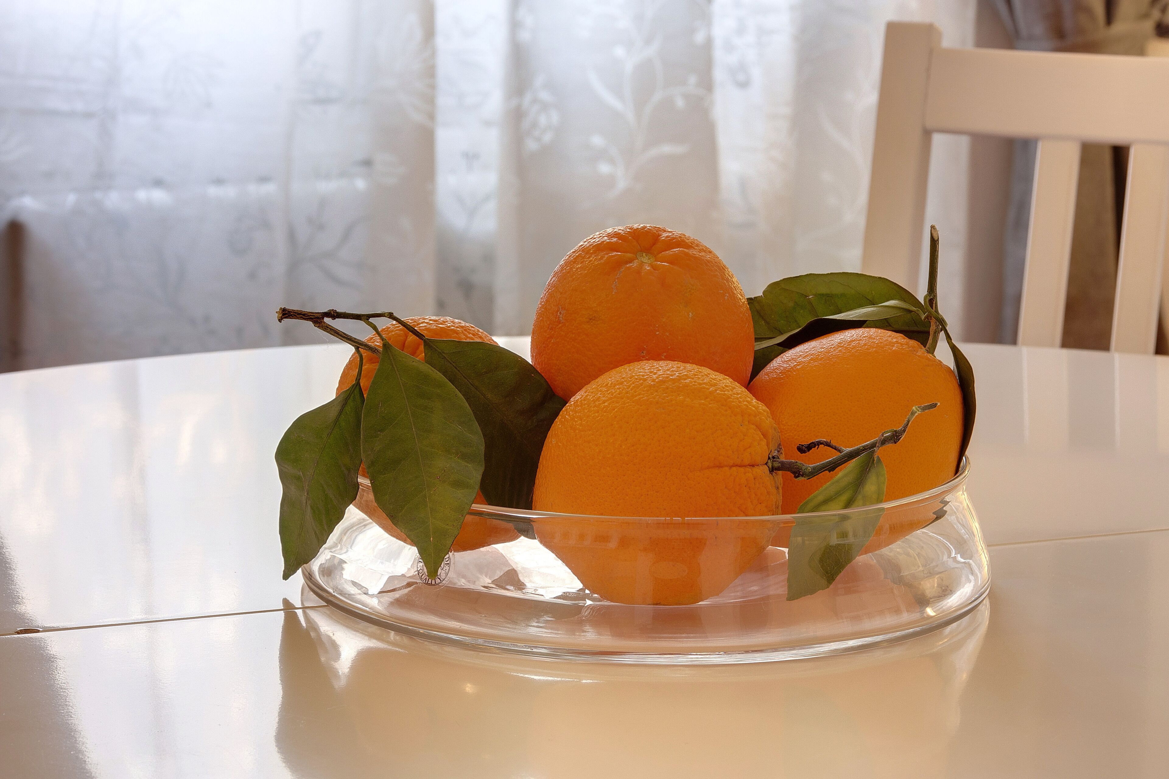 A clear glass bowl with oranges and leaves on a white table.