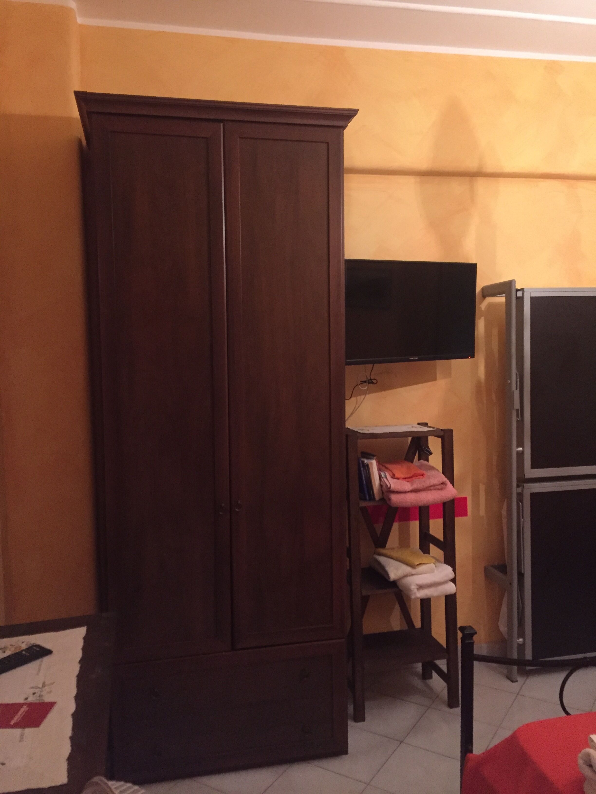 A room with a wooden wardrobe, a television mounted on the wall, a small table with a chair, and a refrigerator.