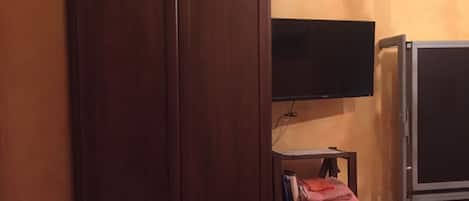 A room with a wooden wardrobe, a television mounted on the wall, a small table with a chair, and a refrigerator.