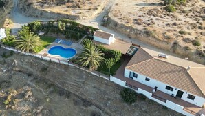 An aerial view of a property with a swimming pool, a white building with a tiled roof, and a surrounding landscape with sparse vegetation. - Villa with sea and mountain views, private pool and large garden (Pitsidia)