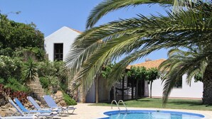 A swimming pool with lounge chairs, surrounded by palm trees and a white building with a tiled roof. - Villa with sea and mountain views, private pool and large garden (Pitsidia)