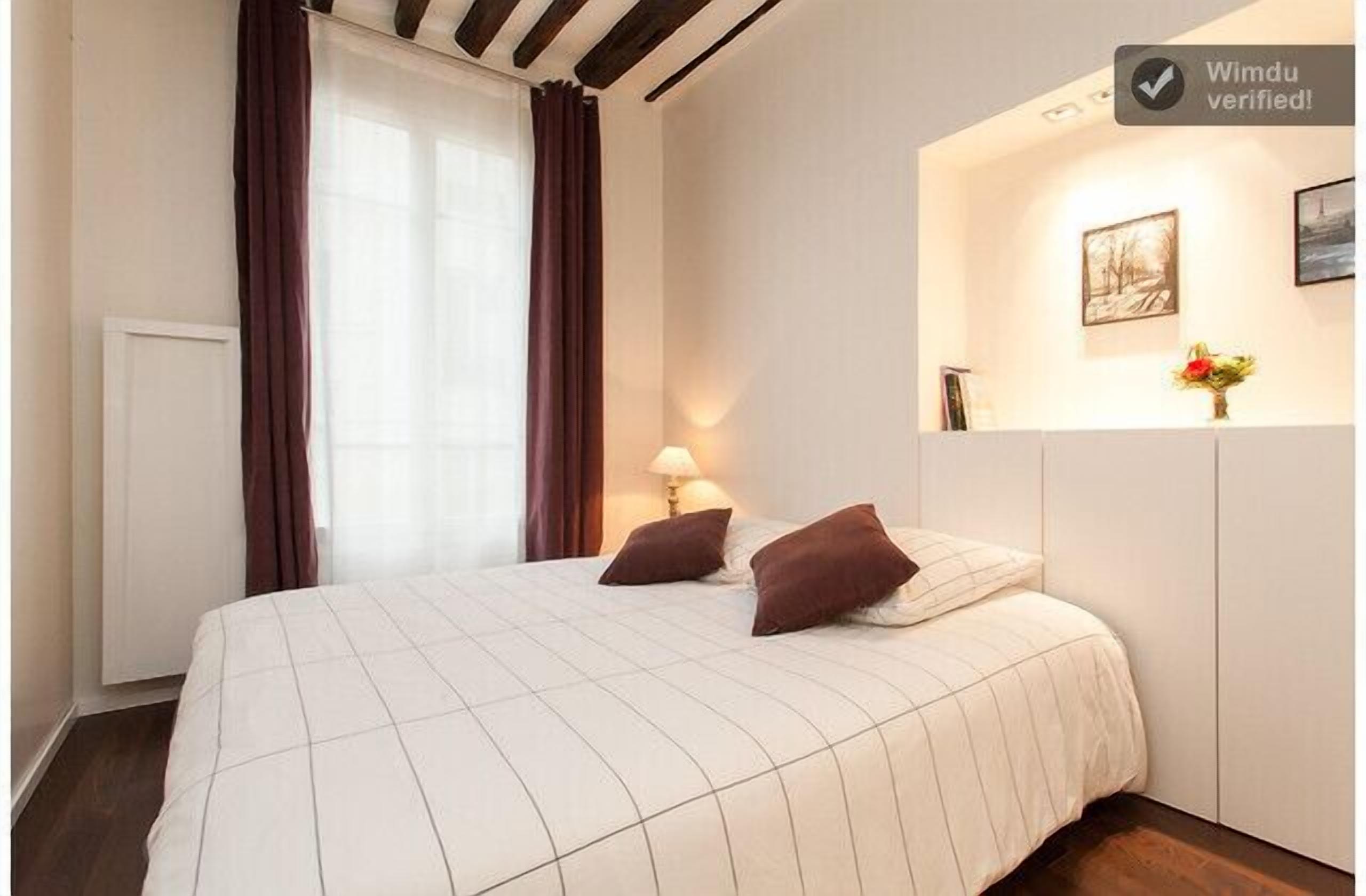 Un dormitorio con cama, dos almohadas, una mesita de noche con lámpara, una ventana con cortinas y un cuadro colgado en la pared.