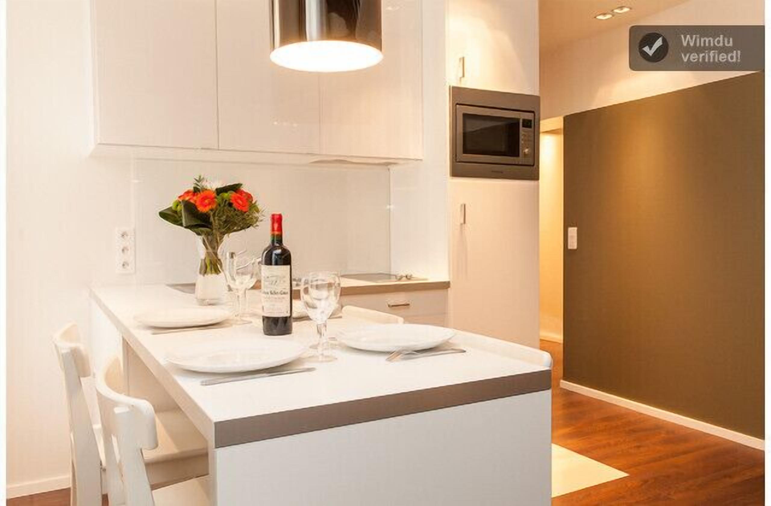 Une cuisine moderne avec des meubles blancs, un four à micro-ondes encastré et un coin repas agrémenté d’une table dressée avec de l’assiette blanche et une bouteille de vin.