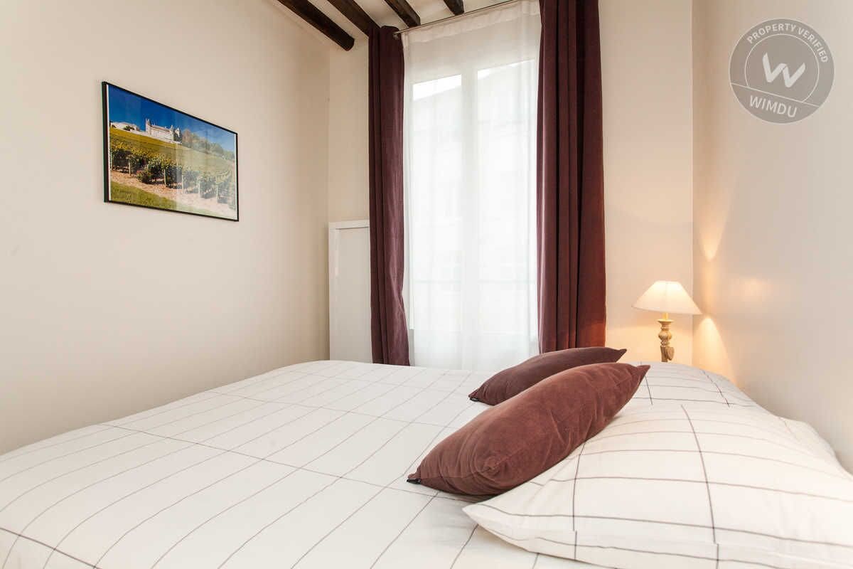 A bedroom with a bed, two pillows, a nightstand with a lamp, a window with curtains, and a wall-mounted picture.