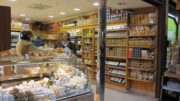 A bakery with a glass display case filled with pastries and bread, shelves stocked with various baked goods and packaged items, and a wooden interior.