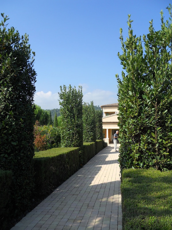 Un chemin pavé mène à un bâtiment dont le toit est recouvert de tuiles, et qui est entouré de haies et de verdure bien entretenues.
