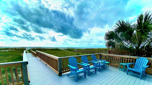 A wooden deck with blue Adirondack chairs and a table, overlooking a beach and grassy dunes. - 9/19-26 WOW SALE! Oceanfront House, 4-7 Night Stay, WEEKLY DEAL, Walk to Pier! (Kure Beach)