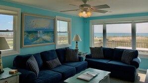 A living room with two dark blue sofas, a ceiling fan, and a large painting of a seascape. - 9/19-26 WOW SALE! Oceanfront House, 4-7 Night Stay, WEEKLY DEAL, Walk to Pier! (Kure Beach)