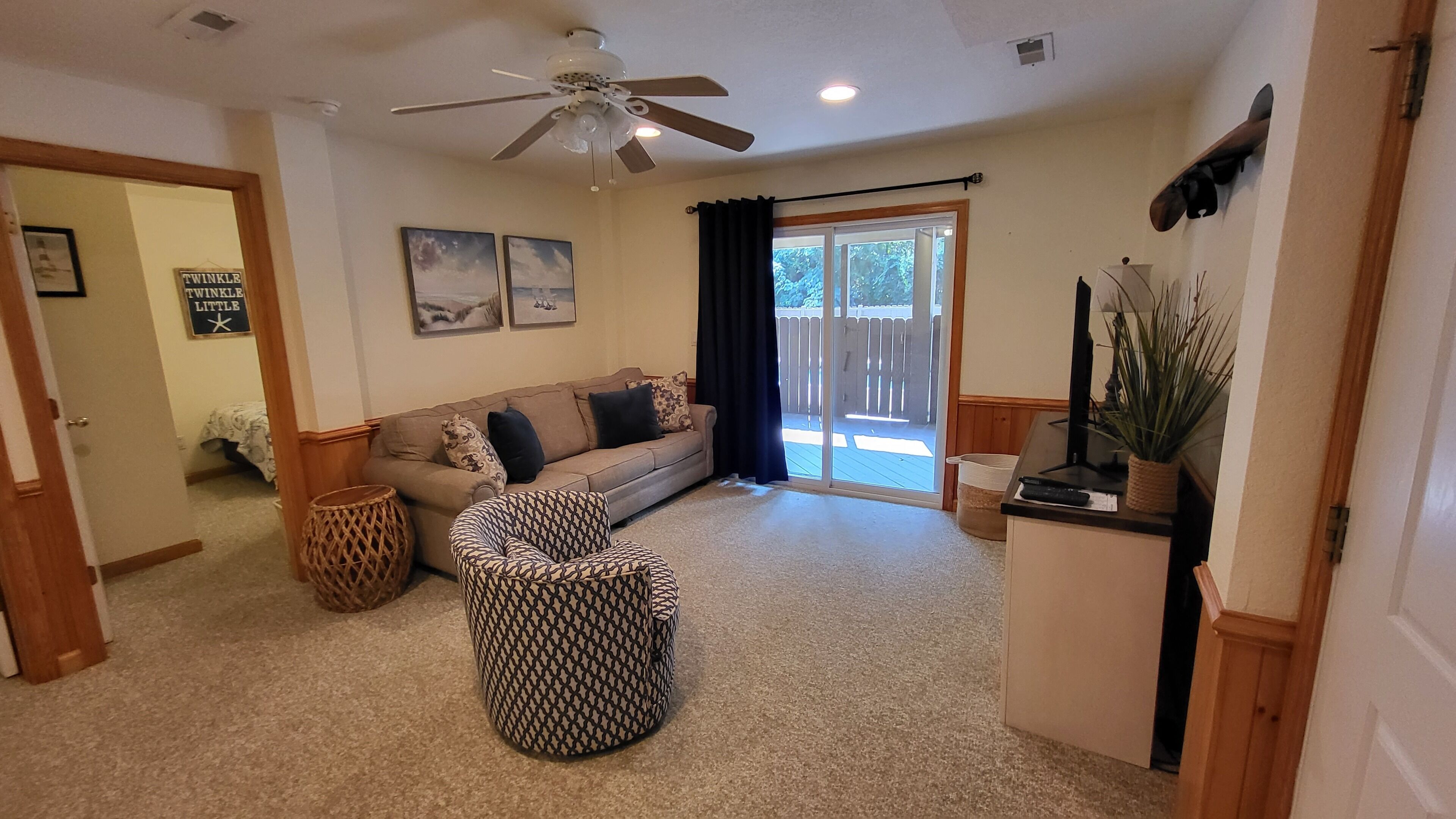 A living room with a beige sofa, patterned ottoman, ceiling fan, and a sliding glass door leading to a patio.