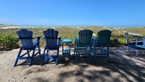 Un área de descanso frente a la playa con sillas Adirondack y una pequeña mesa, con vistas a una playa arenosa y al océano.