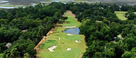 An aerial view of a golf course surrounded by dense forest, with a clear sky and distant water bodies.