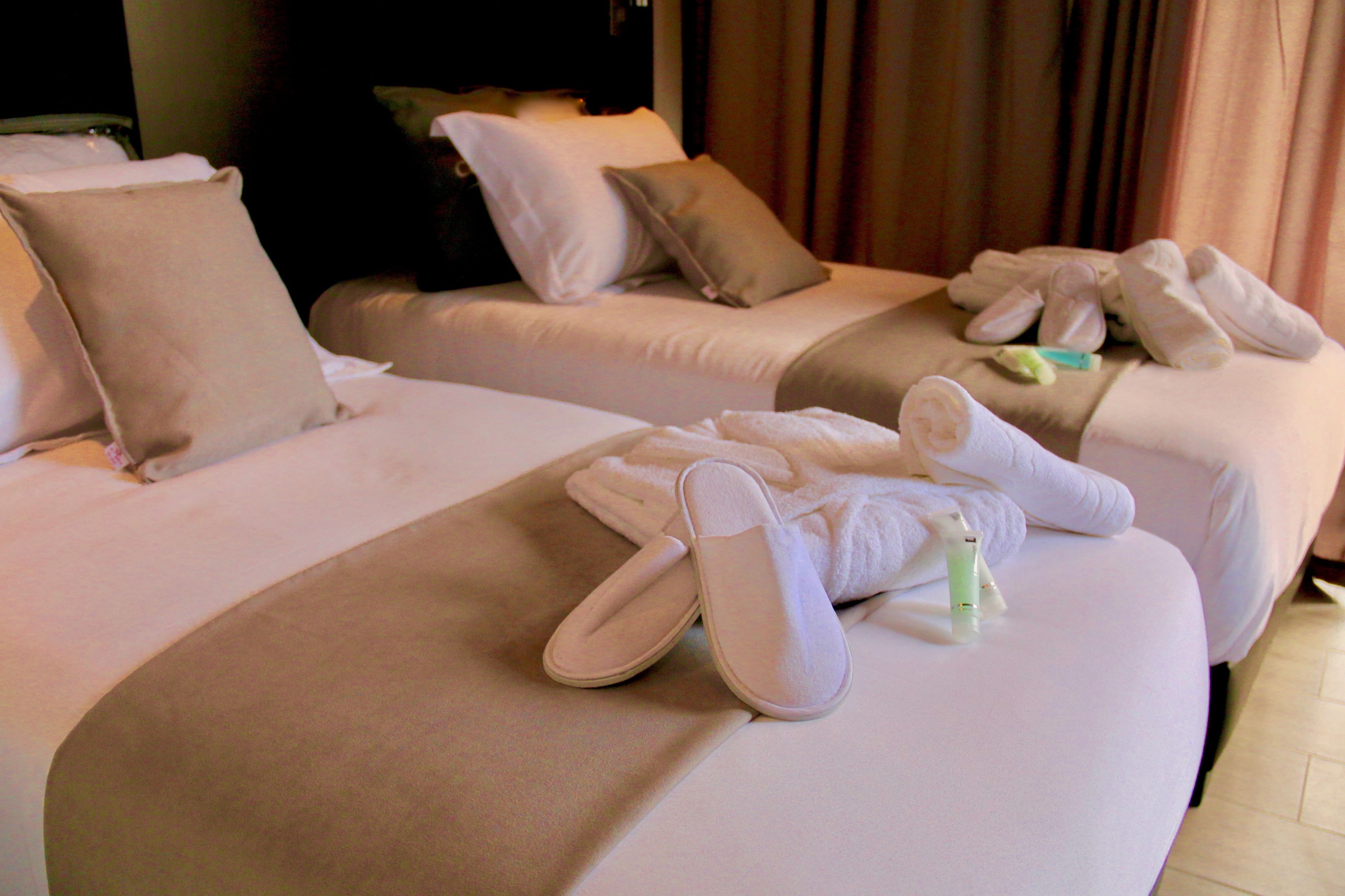 Habitación de hotel con dos camas, cada una con una almohada, una manta y un par de pantuflas.