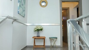Un pasillo con una consola de madera, un jarrón de flores blancas, un espejo redondo y un taburete acolchado verde.