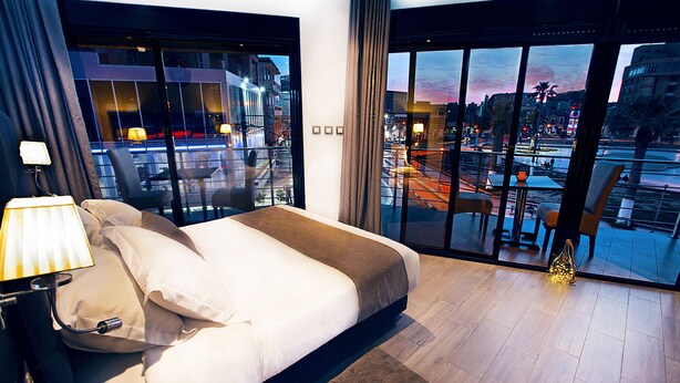 Habitación de hotel moderna con una cama grande, una mesita de noche y vistas a un paisaje urbano a través de ventanales del suelo al techo.