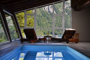 An indoor swimming pool with two wooden loungers and a small table, large windows with a mountain view. - Hotel Puerto Blest (Nahuel Huapi National Park)
