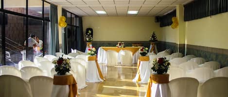 Un salón de banquetes con filas de sillas blancas, mesas con cintas amarillas y centros de mesa florales.