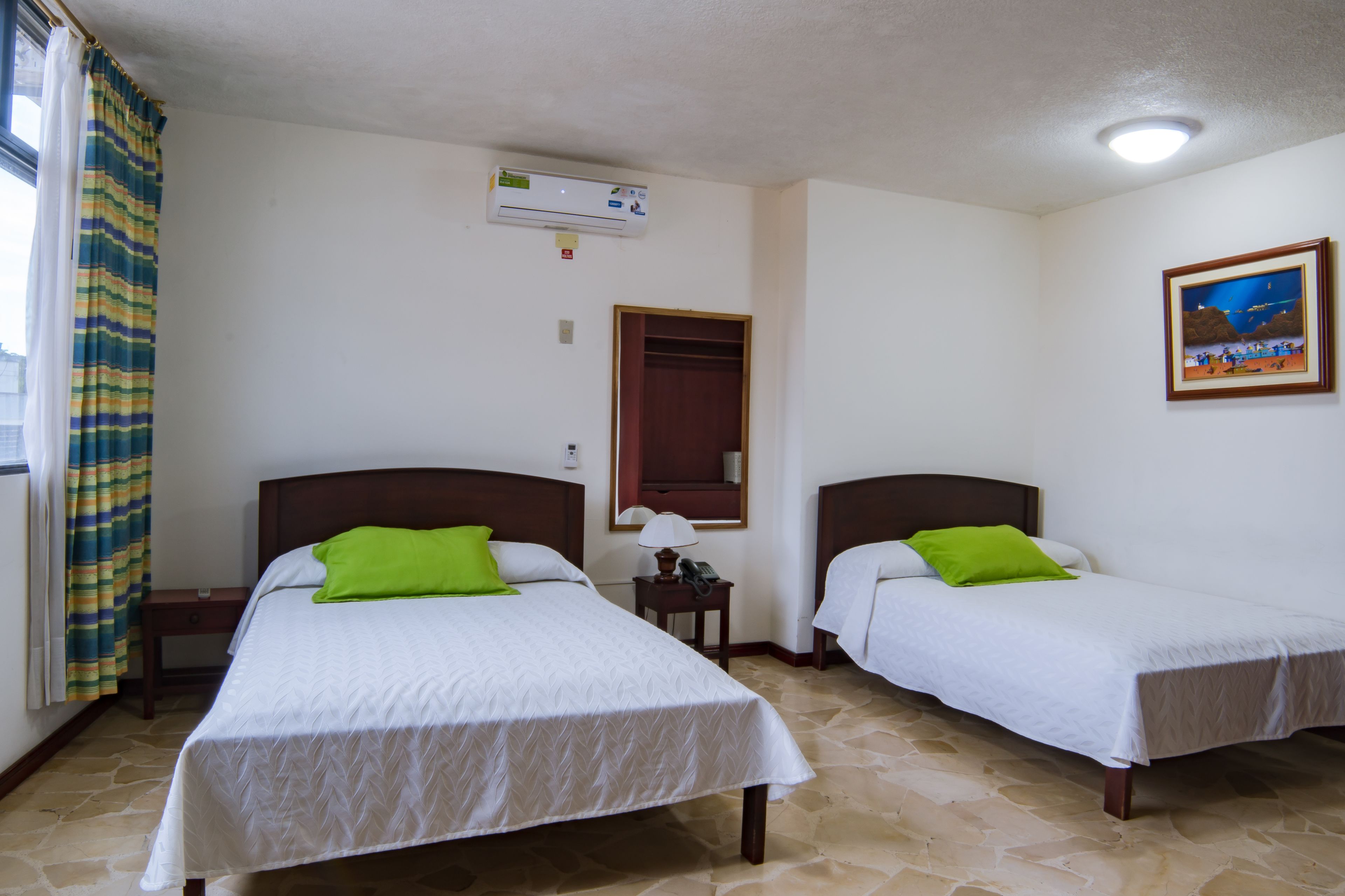 A hotel room with two single beds, a nightstand with a lamp, an air conditioning unit, a framed picture on the wall, and a window with curtains.