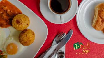 Una escena de desayuno con mantel rojo, que incluye un plato de huevos, dos buñuelos fritos, un tazón de fruta, una taza de café negro y un postre.
